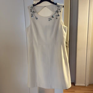 Vit kort klänning med paljettblommor - Superfin vit kort klänning med ärmlös design och snygga paljettblommor vid halsringningen. Klänningen har en markerad midja, dragkedja i ryggen och ett coolt utskuret parti upptill bak. Perfekt för dig som vill sticka ut med eleganta detaljer.
