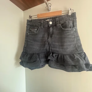 Grå jeanskjol med volang från Gina Tricot - Supersnygg grå jeanskjol från Gina Tricot i kort modell. Kjolen har volang nertill, klassiska jeansfickor fram och bak samt bälteshällor. Perfekt för dig som gillar en cool och trendig stil med extra detaljer.