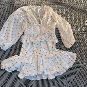 Prickig volangklänning från Shona Joy - Superfin kort volangklänning från Shona Joy i vitt med bruna prickar. Klänningen har långa ärmar, v-ringning, knappar framtill och flera lager volanger. Luftigt material som känns lätt och flowy, perfekt för sommarens alla tillfällen.