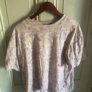 MTV t-shirt med lila tryck - T-shirt i ljusbeige med lila all-over print av MTV-loggor, gitarrer och tecknade figurer. Oversized passform och klassisk rund hals. Perfekt för dig som gillar retrovibbar och streetstyle. Materialet känns mjukt och skönt mot huden.