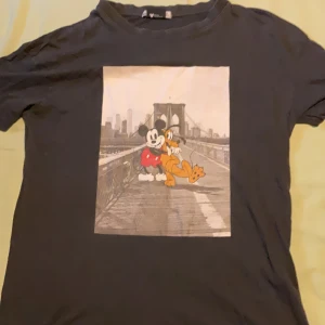 Svart Mickey Mouse t-shirt Pull&Bear - Svart t-shirt från Pull&Bear med stort tryck av Mickey Mouse och Pluto på Brooklyn Bridge. Klassisk rund hals och korta ärmar. Disney-kollektion, perfekt för dig som gillar streetstyle och tecknade motiv.