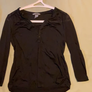 Svart långärmad topp från H&M - Svart långärmad topp från H&M med knappar framtill och lätt v-ringad hals. Tillverkad i mjuk bomullsmix som känns skön mot huden. Perfekt för dig som gillar en enkel och stilren look med lite extra detalj vid halsen. Lite see through.