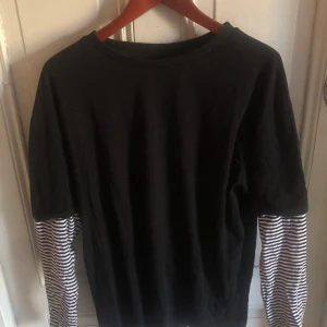 Svart tröja med randiga ärmar - Cool svart tröja med långa ärmar i svartvitt randigt mönster. Tröjan har en avslappnad passform och är gjord i mjuk bomull, perfekt för en chill och stilren look. Passar dig som gillar lager-på-lager-stil.