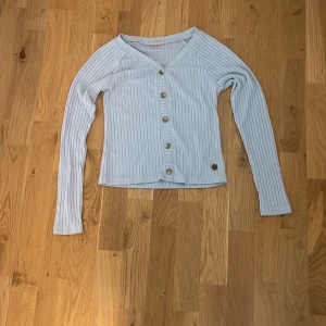 Ljusblå ribbad kofta med knappar - Supersöt ljusblå ribbad kofta från Juicy Couture med v-ringning och snygga pärlemorknappar framtill. Långärmad och i mjuk bomull, perfekt för lager-på-lager. Liten metallbricka med logga nertill ger extra detalj.