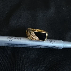 Guldfärgad ring med svart sten - Cool guldfärgad ring med rektangulär svart sten och snedställd rad av små glittrande stenar på ena sidan. Ringen har en chunky känsla och geometriska detaljer på sidorna. Perfekt statement-piece för dig som gillar unika accessoarer.