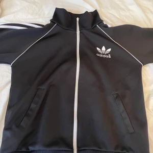 Svart Adidas full zip hoodie med vita detaljer - Svart zip hoodie från Adidas med klassiska vita ränder längs ärmarna och vit piping. Hoodien har dragkedja framtill, hög krage och två fickor. Stort Adidas-tryck på ryggen och mindre logga på bröstet. Perfekt för en sportig och stilren look.