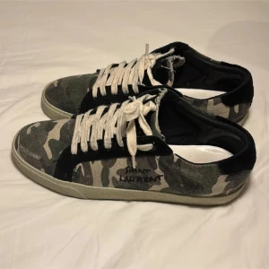 Saint Laurent sneakers i camo - Saint Laurent sneakers med grön och beige camouflage-mönster, svarta detaljer och vita snören. Ovandel i canvas och mocka, platt sula och rund tå. Snygga och unika sneakers som sticker ut i mängden.
