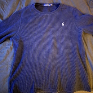 Blå långärmad tröja från Polo Ralph Lauren - Mörkblå långärmad tröja från Polo Ralph Lauren med klassisk rund hals och vit broderad logga på bröstet. Tröjan är i ett strukturerat, stickat bomullsmaterial som ger en avslappnad men ändå stilren vibe. Sitter mer som S i längden men kort över magen