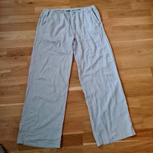 Beiga/grå linnebyxor från 157, storlek M - Luftiga och sköna beiga linnebyxor från 157 med resår och snörning i midjan. Byxorna har raka, vida ben och två sidofickor. Perfekta för varma dagar när du vill ha en avslappnad och chill stil.