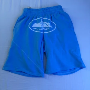 Blå shorts från Corteiz med tryck (Oanvänd) - (Pris kan diskuteras) Snygga blå shorts från Corteiz med elastisk midja och stort vitt tryck framtill. Modellen är loose fit och har en bakficka med knapp. Perfekta för chill dagar eller när du vill sticka ut lite extra. Mjukt material som känns skönt mot huden.
