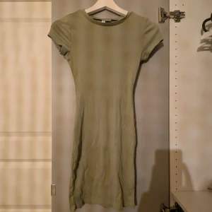 Olivgrön ribbad kortklänning - Supersöt olivgrön kortklänning med ribbad struktur och tight passform. Klänningen har rund halsringning och korta ärmar, perfekt för en enkel och stilren look. Materialet känns mjukt och följsamt mot kroppen.
