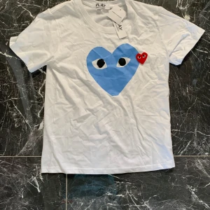 Vit t-shirt Comme des Garçons Play - Vit t-shirt från Comme des Garçons Play med ett stort blått hjärta med ögon och ett litet rött hjärta på bröstet. Klassisk rund halsringning och korta ärmar. Tillverkad i mjuk bomull, perfekt för en clean och cool look.