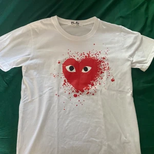 Vit Comme des Garçons Play t-shirt - Vit t-shirt från Comme des Garçons Play med ikoniskt rött hjärta med ögon och färgstänk på bröstet. Klassisk rund hals och korta ärmar. Perfekt statement-plagg för dig som gillar streetstyle och unika prints.