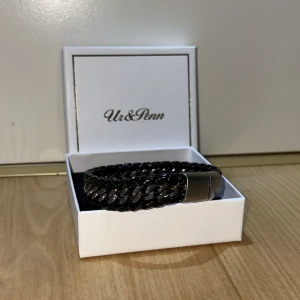 Svart flätat armband Ur & Penn - Stilrent svart armband från Ur & Penn med flätad design och blanka silverfärgade detaljer. Armbandet har ett coolt kedjemönster och magnetlås i metall som ger en modern vibe. Perfekt accessoar för att lyfta din outfit.