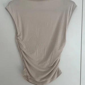 Beige ärmlös topp från Gina Tricot - Snygg beige ärmlös topp från Gina Tricot i mjukt och stretchigt material. Toppen har drapering på ena sidan som ger en cool och trendig look. Perfekt att styla med jeans eller kjol för en fräsch outfit.