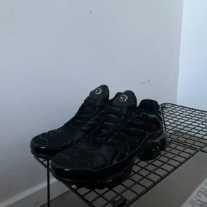 Svarta Nike Air Max Plus TN sneakers - Svarta Nike Air Max Plus TN sneakers med helsvart design och diskreta vågformade detaljer på ovandelen. Skorna har mesh och syntetmaterial, Tn-logga bak och klassisk platt sula. Perfekta för dig som gillar en clean och sportig look. med kvitto