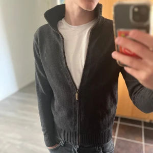 Svart kofta från Polo Ralph Lauren - Säljer en mörk grå stickad kofta från Polo Ralph Lauren med hel dragkedja och hög krage. Tröjan har ribbade muddar och nederkant, samt långärmad passform. Perfekt för dig som gillar klassisk och stilren look. Perfekt till sommarkvällar, mycket bra skick storlek 14-16 så passar xs
