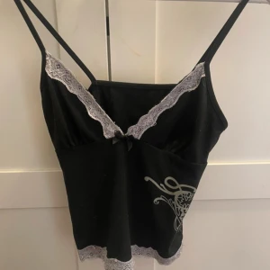 Svart linnetopp med spets och tryck - Snygg svart linnetopp med tunna axelband, vit spetskant vid ringning och nederkant samt ett coolt grått tryck på sidan. Toppen har en liten rosett framtill och v-ringad urringning. Perfekt för dig som gillar en trendig och feminin stil.