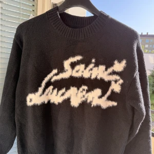 Svart Saint Laurent stickad tröja - Svart stickad tröja från Saint Laurent med vit, fluffig logotyp framtill. Tröjan har rund halsringning, långa ärmar och ribbade muddar. Perfekt för dig som vill ha en statement piece med lyxig känsla och cool kontrast mellan svart och vitt.