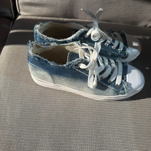 Blå jeanssneakers med fransad kant - Snygga sneakers i blått jeanstyg med fransade kanter och vita snören. Skorna har vit tå och sula, vilket ger en cool kontrast till denimlooken. Perfekta för dig som gillar en avslappnad och trendig stil.