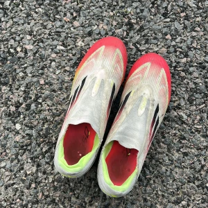 Adidas F50 Elite - Snabba Adidas X Speedflow fotbollsskor i rött, vitt och neongult. Skorna har en strömlinjeformad design utan snörning, med svarta Adidas-ränder på sidorna och lätt syntetmaterial för maxad fart på planen.