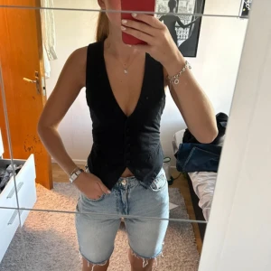 Svart väst med knappar från H&M - Snygg svart väst från H&M med djup v-ringning och knäppning framtill. Västen har en figurnära passform och justerbart spänne i ryggen. Passar en 36 också.