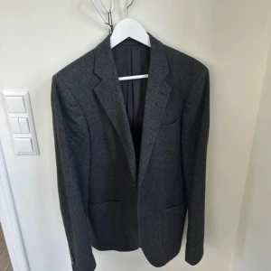 Mörkgrå kavaj från Tiger of Sweden - Stilren mörkgrå kavaj från Tiger of Sweden med klassisk passform och snyggt snitt. Kavajen har två framfickor, knappar vid ärmslut och är helfodrad. Perfekt för dig som vill ha en tidlös och elegant look.