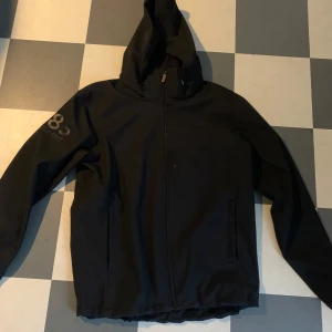 Svart vindjacka med huva Didriksons - Svart vindjacka från Didriksons med huva och hel dragkedja framtill. Jackan har diskret logga på bröstet och tryck på ena ärmen. Perfekt för blåsiga dagar och enkel att matcha med allt. Snygg och funktionell design med två fickor framtill.