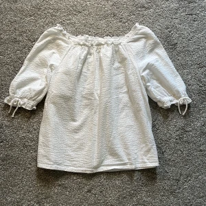 Vit offshoulder blus från H&M - Superfin vit offshoulder blus från H&M i storlek 164. Blusen har trekvartsärmar med knyt vid ärmsluten och en lätt strukturerad bomullskänsla. Söt volangdetalj längs halsringningen och luftig passform. 