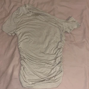 Beige oneshoulder topp Gina Tricot XS - Snygg beige topp från Gina Tricot i oneshoulder-modell. Toppen har kort ärm på ena sidan och rynkade detaljer längs sidorna som ger en cool och trendig look. Perfekt för dig som gillar att sticka ut lite extra. Står xs men efter att jag har tvättat och de så känns det faktiskt som att de passar xxs osså