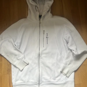 Vit hoodie från Sail Racing - Säljer en stilren vit hoodie från Sail Racing med dragkedja framtill och logga på bröstet. Tröjan har huva med snörning, ribbade muddar och en praktisk bröstficka med dragkedja. Perfekt för dig som gillar sportig och clean stil.