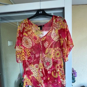 Blommig blus från H&M i mesh - Blus från H&M i transparent mesh med blommigt mönster i rött, rosa och gult. Modellen har v-ringning, korta ärmar och en lätt, luftig känsla. Storlek 34.