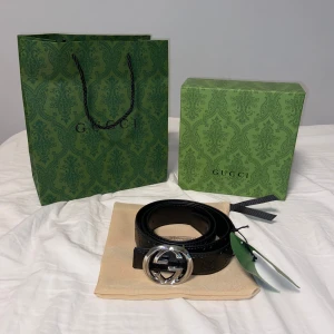 Svart bälte med silver spänne - Snyggt svart bälte i skinn med präglade loggor längs hela bandet. Bältet har ett stort, rundat spänne i silverfärgad metall. Perfekt accessoar för att lyfta vilken outfit som helst.