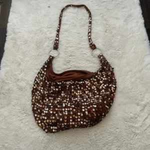 Brun axelväska med paljetter Fashion Express - Snygg brun axelväska från Fashion Express med glittriga paljetter över hela väskan. Väskan har en mjuk form, bred axelrem med silvriga ringar och dragkedja upptill. Perfekt för dig som vill sticka ut med en unik accessoar.