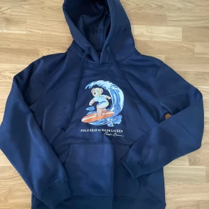 Mörkblå Polo Bear hoodie från Ralph Lauren - Säljer en mörkblå hoodie från Ralph Lauren med den ikoniska Polo Bear som surfar på framsidan. Tröjan har huva med snörning och en stor magficka. Mjuk bomullskänsla och klassisk passform, perfekt för dig som gillar streetstyle och snygga prints. Den har 2 defekter vilket är på snörerna som ni ser i bild 3, alltså att dem gått av där, pga av att jag råka göra sönder. Men annars är tröjan helt ny som ni ser på bilderna👍
