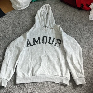 Grå hoodie med tryck från Gina Tricot - Säljer en ljusgrå hoodie från Gina Tricot med svart text 'AMOUR' på framsidan och en stjärna på ryggen. Tröjan har huva, ribbade muddar och är tillverkad i mjuk bomullsblandning. Perfekt för chill dagar eller när du vill ha en enkel men snygg look.