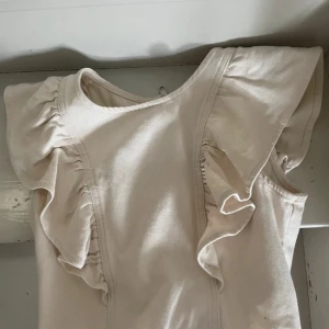 Beige blus med volangärmar - Supersöt beige blus med volangdetaljer på axlarna och dragkedja i ryggen. Ärmlös modell med rund halsringning och mjukt bomullstyg som känns skönt mot huden. Perfekt för dig som gillar en stilren men ändå lekfull look.