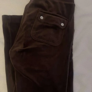 Bruna juicy byxor - Mörkbruna mjukisbyxor i mjuk sammet med bred resår i midjan och stora fickor med tryckknappar. Snygg detalj med broderad logga på ena fickan. Perfekta för en chill och avslappnad stil. 