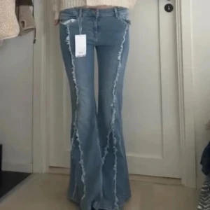 Blå flare jeans med fransade detaljer - Säljer ett par blåa flare jeans från Bershka med råa, fransade sömmar längs benen. Jeansen har hög midja och en riktigt snygg utsvängd passform som ger retrovibbar. Perfekta för dig som vill sticka ut med coola detaljer och trendig look.