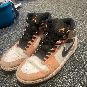 Nike Air Jordan 1 Mid i beige /gul och svart - Snygga Nike Air Jordan 1 Mid med beige mockadetaljer, vita partier och svarta detaljer som swoosh, snörning och plös. Guldigt Jumpman-logo på plösen. Klassisk high-top siluett med rund tå och platt sula. Perfekta för dig som vill sticka ut med en ikonisk sneaker.
