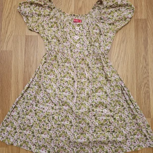 Blommig kortklänning från m.m.f.l. - Supersöt kortklänning med blommigt mönster i rosa, grönt och vitt. Klänningen har korta ärmar, knappar framtill, snörning i midjan och två fickor med knappdetaljer. Perfekt för sommarens alla tillfällen!