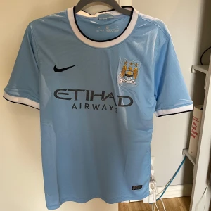Manchester City Kompany #4 Nike tröja - Manchester Citys officiella matchtröja med Kompany och nummer 4 på ryggen. Ljusblå med vita och svarta detaljer vid krage och ärmslut, broderat klubbmärke och Nike-logga. Tillverkad i lätt, ventilerande Dri-FIT-material. Sponsortryck från Etihad Airways på bröstet.