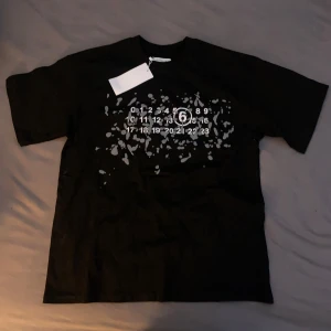 Svart t-shirt Maison Margiela splatter - Svart t-shirt med ikoniskt siffror-tryck och grått splattermönster på bröstet. Klassisk passform med korta ärmar och rund hals. Tillverkad i mjuk bomull för en skön känsla. Perfekt för dig som gillar streetstyle och unika designer.