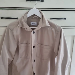 Beige overshirt från Reserved Eco Aware - Stilren beige overshirt från Reserved Eco Aware med två bröstfickor och svarta knappar framtill. Skjortan har klassisk krage och långa ärmar med knapp vid ärmslut. Perfekt att bära öppen eller stängd för en avslappnad look.