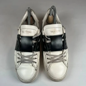 Valentino Open - Snygga Valentino Garavani Open sneakers i vitt skinn med bred svart läderdetalj över snörningen. Grå skosnören och ikoniska nitar baktill på sulan. Klassisk rund tå och platt sula, perfekt för en trendig streetstyle-look.