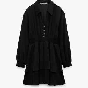 Svart volangklänning från Zara - Supersnygg svart kortklänning från Zara med volanger, långärmad design och markerad midja. Klänningen har v-ringning med knappar framtill och en klassisk krage. Perfekt för dig som gillar en stilren och trendig look.