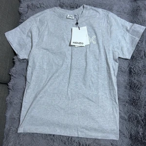 Grå t-shirt från Kenzo med logga - Snygg grå t-shirt från Kenzo med vit logga tryckt på bröstet. Klassisk rund halsringning och korta ärmar. Tillverkad i mjuk bomull med lite elastan för extra komfort. Perfekt för en clean och stilren look.