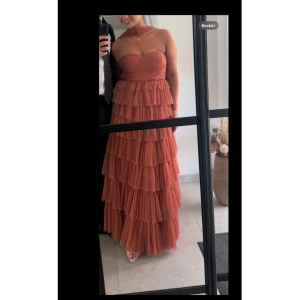 Festklänning/ volangklänning med bara axlar och scarf  - Säljer en lång rostbrun klänning med flera lager volanger och bara axlar. Klänningen har omlottliv och en snygg scarf-detalj runt halsen. Perfekt för bal eller fest, ger en riktigt elegant och trendig look.