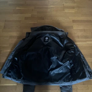 Svart pufferjacka från Canada Goose - Säljer en grå pufferjacka från Canada Goose med klassisk logga på ärmen. Jackan är vadderad och har en hög krage, perfekt för kalla dagar. Tillverkad i slitstarkt material med mjukt foder och ribbade muddar vid ärmsluten. Snygg och tidlös modell som håller dig varm. 