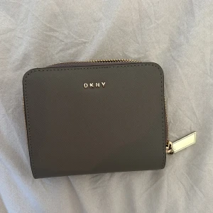 Grå plånbok från DKNY - Plånbok från DKNY i stilren design med guldfärgade detaljer och dragkedja. Plånboken har flera kortfack, myntfack och går att stänga med både knapp och dragkedja. Använd typ en gång så helt ny🥰